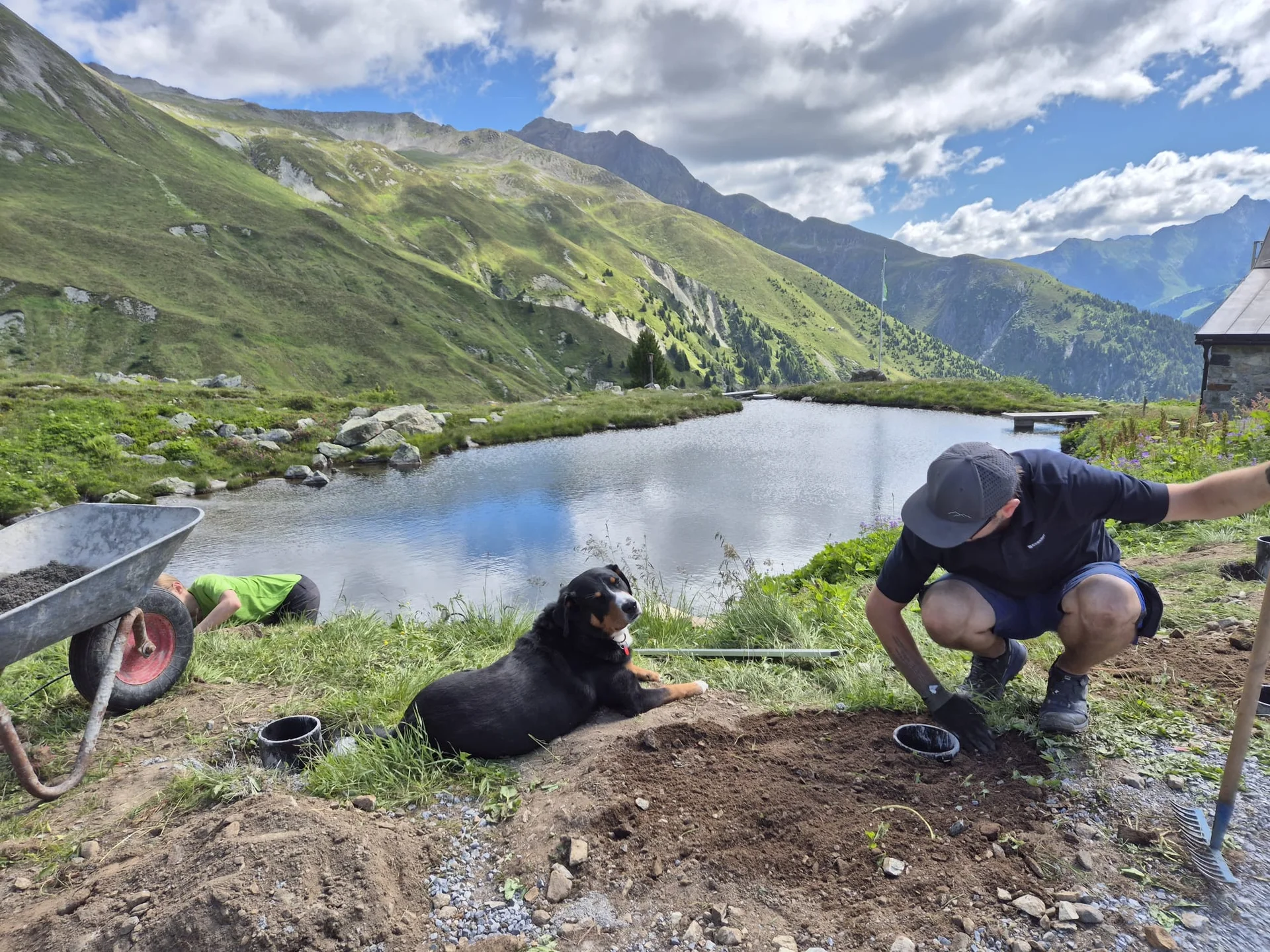 Mann kniet neben dem See an der Häfler Hütte, neben ihm liegt ein schwarzer Hund, im Hintergrund grüne Hügel und blauer Himmel | © DAV-FN / R. Klees o. R. Zahner