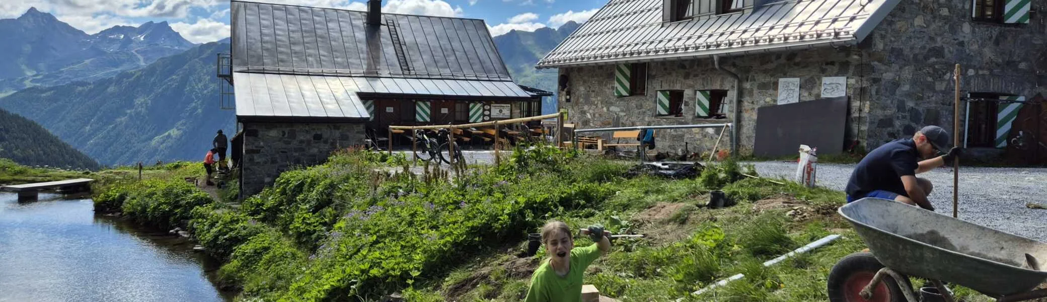 Zwei Personen graben auf einer Wiese neben dem Bergsee, im Hintergrund die Häfler Hütte und Berge unter blauem Himmel mit Wolken | © DAV-FN / R. Klees o. R. Zahner