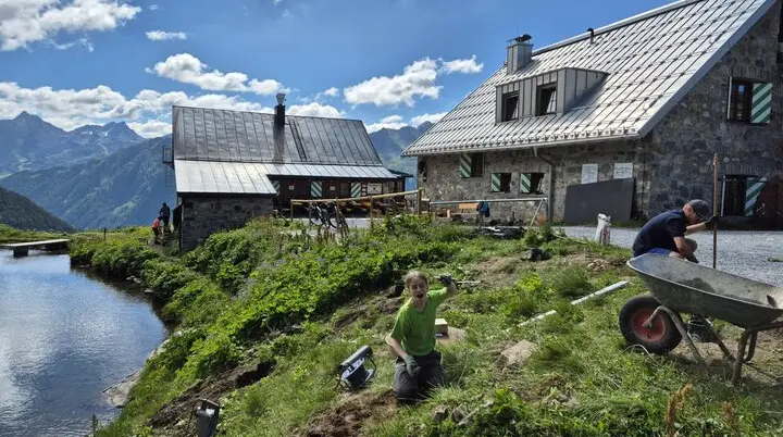 Zwei Personen graben auf einer Wiese neben dem Bergsee, im Hintergrund die Häfler Hütte und Berge unter blauem Himmel mit Wolken | © DAV-FN / R. Klees o. R. Zahner