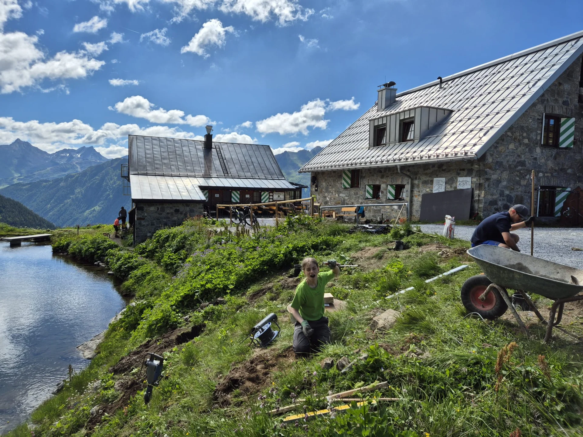 Zwei Personen graben auf einer Wiese neben dem Bergsee, im Hintergrund die Häfler Hütte und Berge unter blauem Himmel mit Wolken | © DAV-FN / R. Klees o. R. Zahner